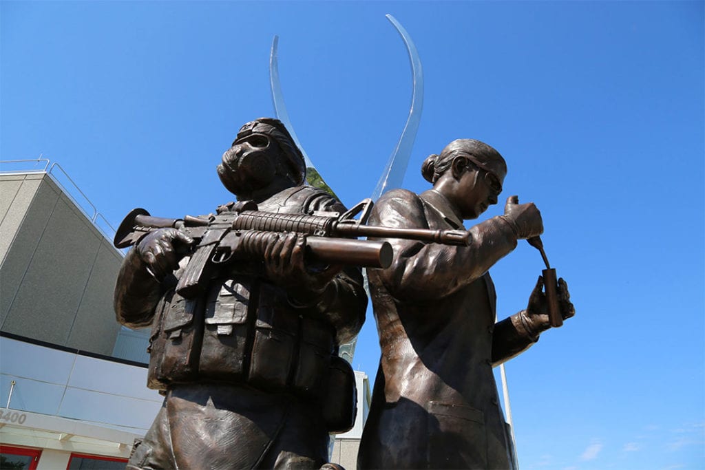 Bronze statue of solider in gas mask standing back to back with a scientist pipetting into a test tube. Statue is outside building against a backdrop of blue sky.