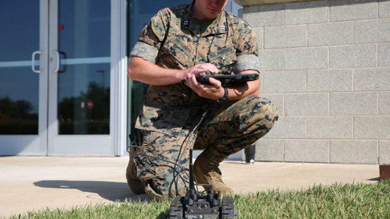 Marine in camouflage uniform kneels cradling controller board to operate small robot in front of him in the grass