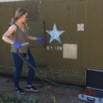 Gloved chemist stands outside large green metal army bunker with hose-like device attached to sensor in metal case