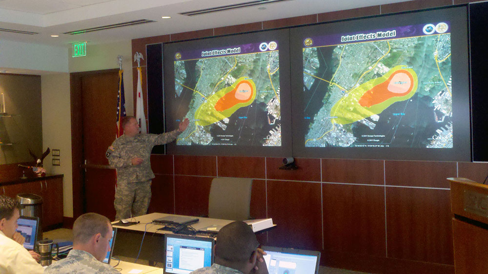 Dual screens behind presenter showing large area maps with orb plume indicating contaminated areas