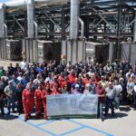 In front of large industrial structure with gleaming metal pipework, dozens of workers stand with a banner that marks the date and says 'Last Munition Destroyed'.