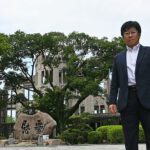 A large multi-story stone structure obscured by mature trees. In front of this is a serious-looking academic dressing in a suit with no tie, looking directly at the camera.