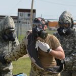 two military personnel in chem gear help a third person remove simulated contaminated clothing. All are wearing gas masks.