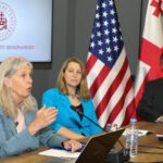 three people gathered at a table in thoughtful discussion with the American and Georgian flags displayed behind them.