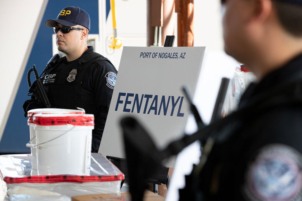 Two U.S. Customs and Border Protection officers in tactical gear stand near a large white container and a sign labeled "Fentanyl" at the Port of Nogales, Arizona, highlighting the growing need to protect first responders from fentanyl exposure.