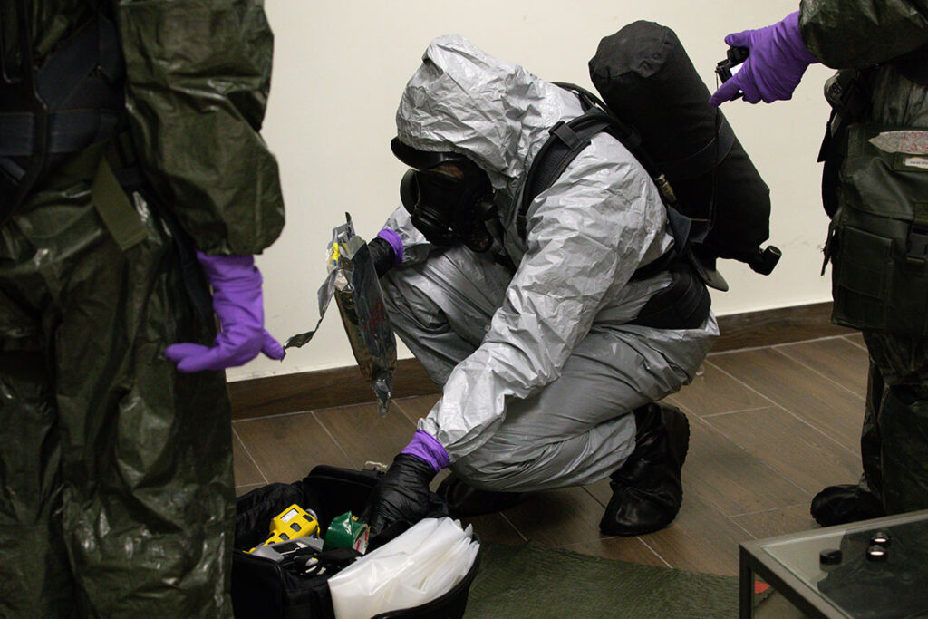A person in a fully encapsulated protective suit with an air tank and full-face respirator is crouched on one knee indoors, examining a sealed sample bag from an open equipment case containing radiation detection instruments and supplies. Two other similarly equipped individuals stand nearby, one pointing toward the sample. The scene depicts a controlled training environment for responding to radiological threats, emphasizing hands-on practice with contamination detection and evidence handling.