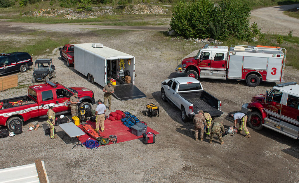 Emergency responders, including military personnel and firefighters, organize gear from multiple trucks and trailers in a gravel staging area during a joint disaster response exercise involving simulated casualty scenarios. Equipment includes stretchers, packs, generators, and medical supplies.