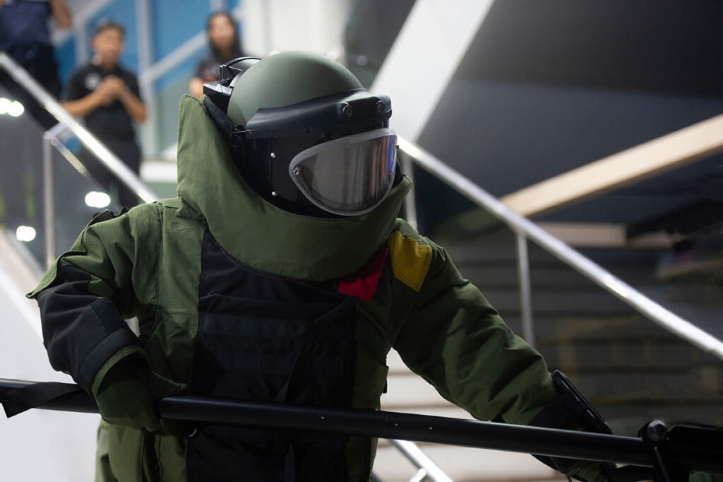 A bomb disposal technician wearing a full-body protective suit and helmet visor stands indoors near a staircase, holding a tool used for handling explosive devices. The person is engaged in an explosive ordnance disposal response scenario in a modern urban environment.