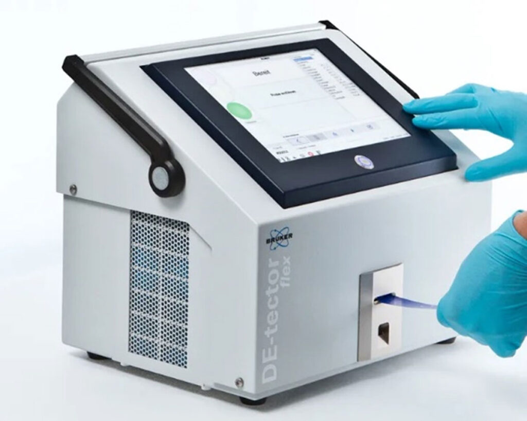 A gloved hand inserts a sample swab into the Bruker DE-tector flex device, a tabletop system used to detect trace levels of explosives and chemicals in security screening environments. The device has a touchscreen interface and is in active use.