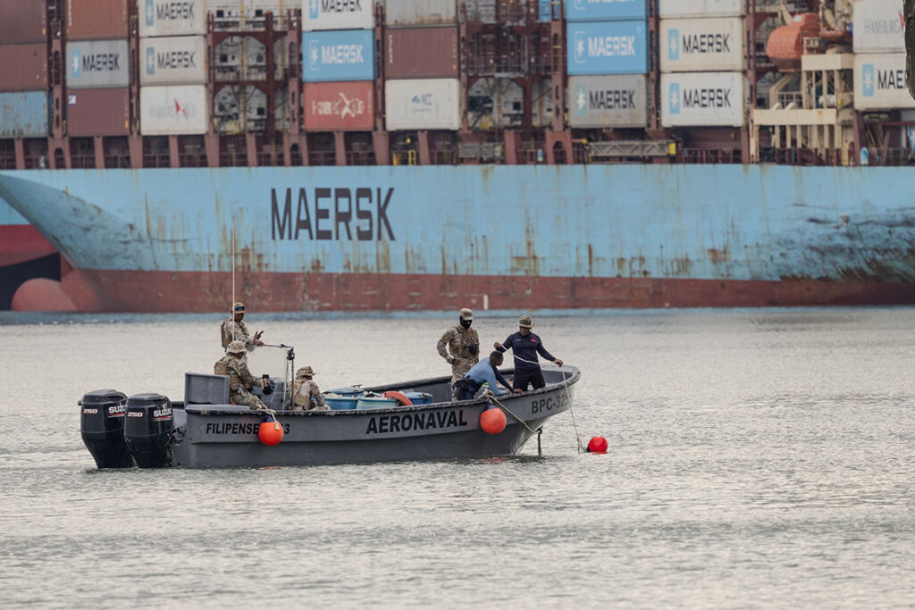 A small security boat labeled "AERONAVAL" with several uniformed personnel aboard conducts underwater inspection near floating markers. In the background, a large Maersk container ship is visible, indicating proximity to a major maritime shipping route. The personnel are engaged in maritime security training to detect and neutralize explosive threats in a high-traffic waterway.
