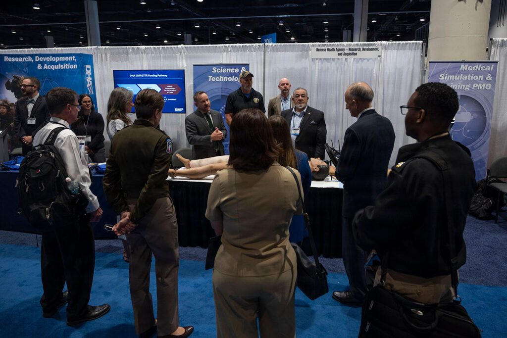 Defense Health Agency senior leaders and military medical professionals gather at an exhibition booth during the symposium. A presenter speaks to the group while displaying information about research, development, and acquisition initiatives. Multiple attendees stand around a table with documentation and displays. Booth signage shows organizational branding and research program information. The professional conference setting shows other exhibition booths and institutional displays in the background.
