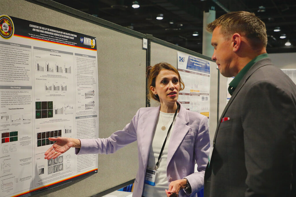 A researcher in professional attire stands at a research poster presentation, gesturing toward scientific data displayed on the poster board. A military attendee faces the presenter and listens to the research explanation. The poster displays multiple sections including introduction, key findings, and functional alterations, with charts, graphs, and microscopy images visible. The symposium setting shows institutional branding and other research displays in the background.