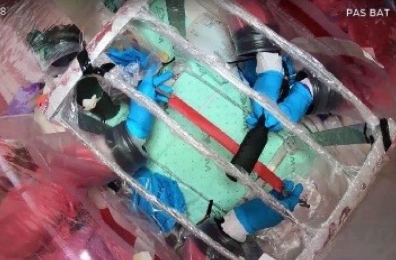 An overhead view inside a transparent, inflated glove bag containing a light green rectangular surface with strips of dark and red tape laid across it in a crosshatch pattern. Several pairs of black and blue gloves, some built into the bag's ports and others loose, are positioned around the edges of the workspace. Additional bundled materials and packaging are visible around the perimeter of the bag. Onscreen video overlay text in the top right corner reads "PAS BAT."
