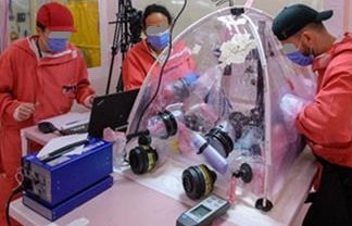 Three people in red protective suits, masks, and gloves work at a table with a large transparent inflated glove bag containing multiple cameras on tripods pointed at evidence inside. A blue light source unit sits on the table to the left, connected by a cable running toward the bag. Additional equipment, including a laptop and handheld device, sits on the tabletop.