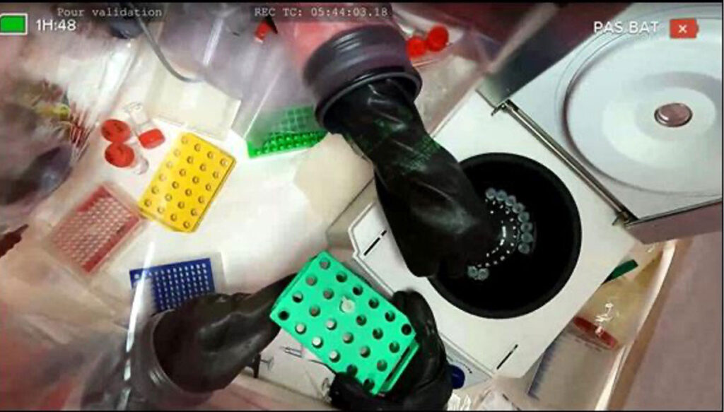 Forensic Teams Learn to Solve Crimes at Radioactive Scenes Without Getting Contaminated An overhead view from inside a transparent glove bag shows two gloved hands manipulating laboratory equipment. One hand lowers a rack of small sample vials into a circular centrifuge well, while the other holds a green multi-well sample tray. Surrounding the workspace are additional colored sample trays in yellow, red, and blue, along with small vials and a disc-shaped piece of equipment. Onscreen video overlay text at the top reads "Pour validation," a timecode, and "PAS BAT" with a red icon.