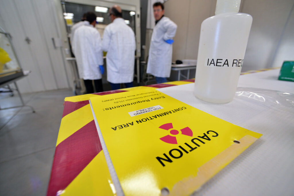 In the foreground, a yellow warning sign with a radiation trefoil symbol reads "CAUTION CONTAMINATION AREA" alongside entry requirement text, positioned on a table next to a labeled sample bottle marked "IAEA REC..." In the background, three people wearing white lab coats and blue gloves stand and converse near a fume hood and workbench inside a laboratory space.